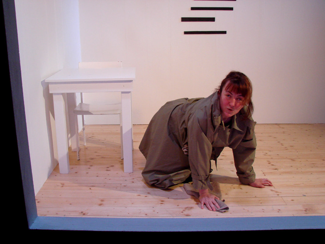 Fotografija: Na tleh kleči igralka, naslonjena na roki, v desni drži krpo s katero briše tla. 