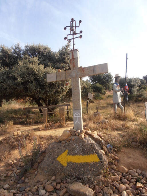 Fotografija: Na nasutem kamenju stoji križ, pod katerim je rumena smerna puščica, ki kaže v levo. Za križem se opazi spomenik pohodnika, v rokah drži palico in čevlje.