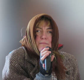 Fotografija: Na videz ostarela ženska, na glavi ima rjavo ruto, na rokah strgane rokavice, v rokah pa drži mikrofon. 