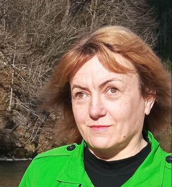Fotografija:  Obraz nasmejane Metke. Ima svetle lase, ki ji segajo do ramen, rahlo naličene ustnice, rjavkasto zelene oči, oblečen ima živo zelen plašč pod katerim nosi črn puli. V ozadju reka Idrijca in drevesa.