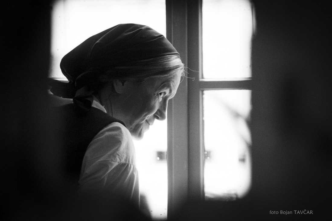Fotografija: Obraz igralke v vlogi Marine Melhiorce, ki nagajivo pogleduje skozi majhno okence. Na glavi ima zadaj zavezano temno ruto izpod katere se vidi pramen sivih las.